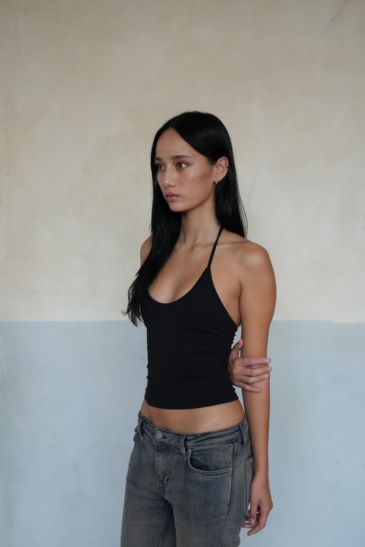 Noa in a black top and jeans against a two-tone plaster wall